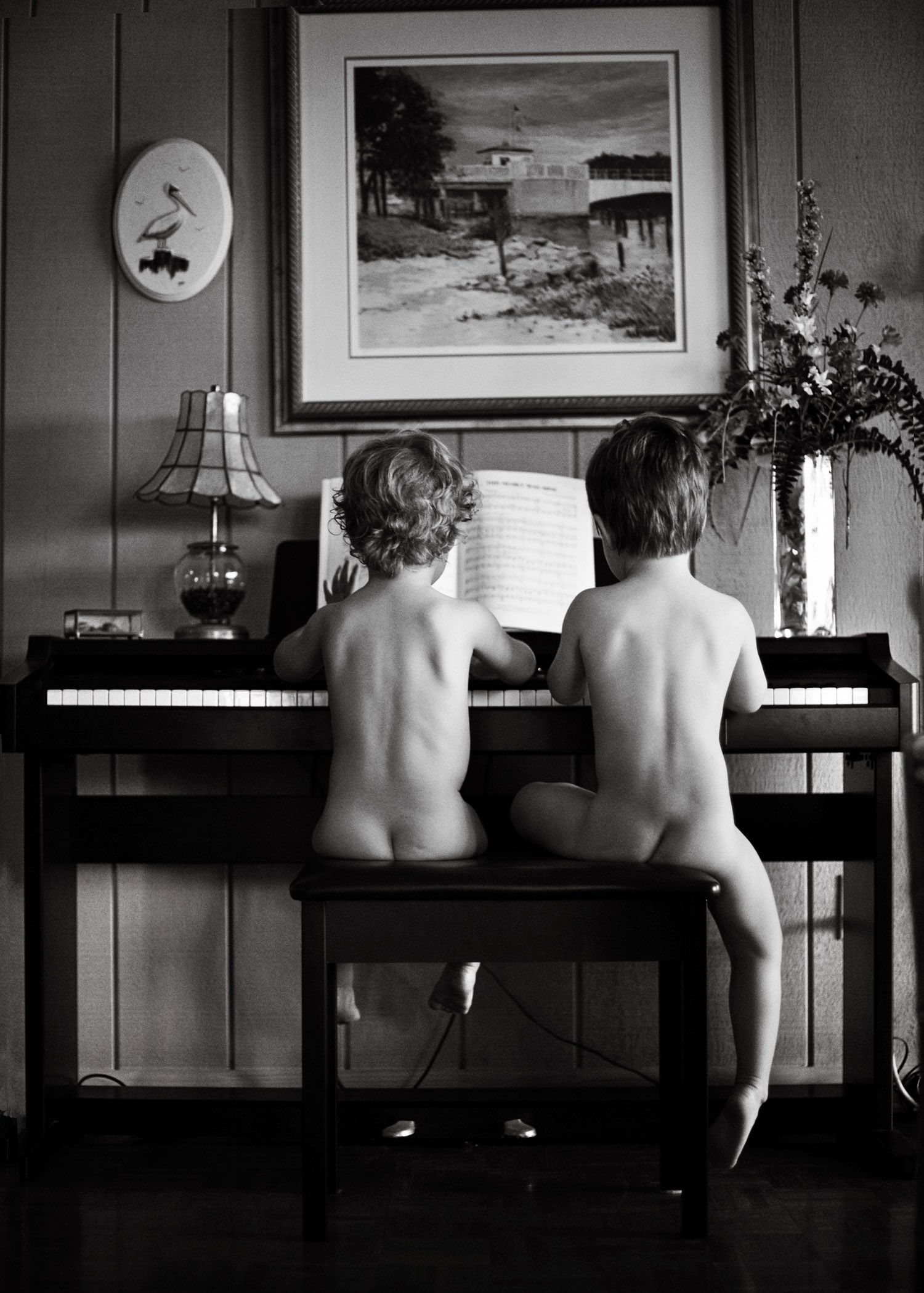 A black and white vintage photograph shows two small children seated at a piano with framed artwork above.