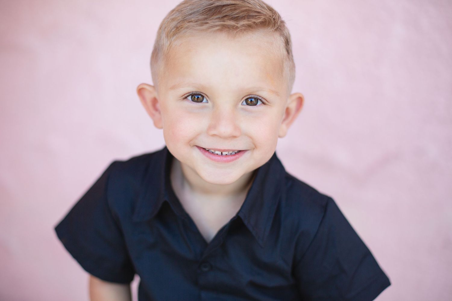 A young child with a bright smile wearing a navy blue polo shirt against a pink background.