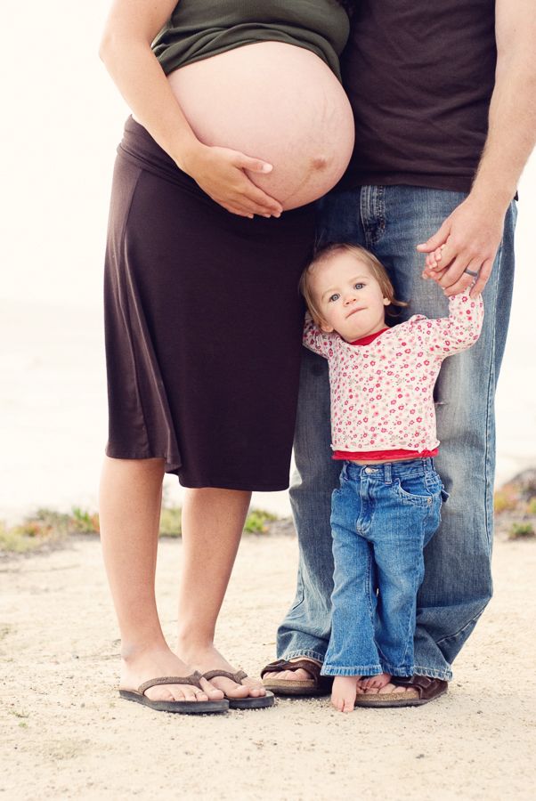 Toddler stands between two figures showing pregnant belly at beach.
