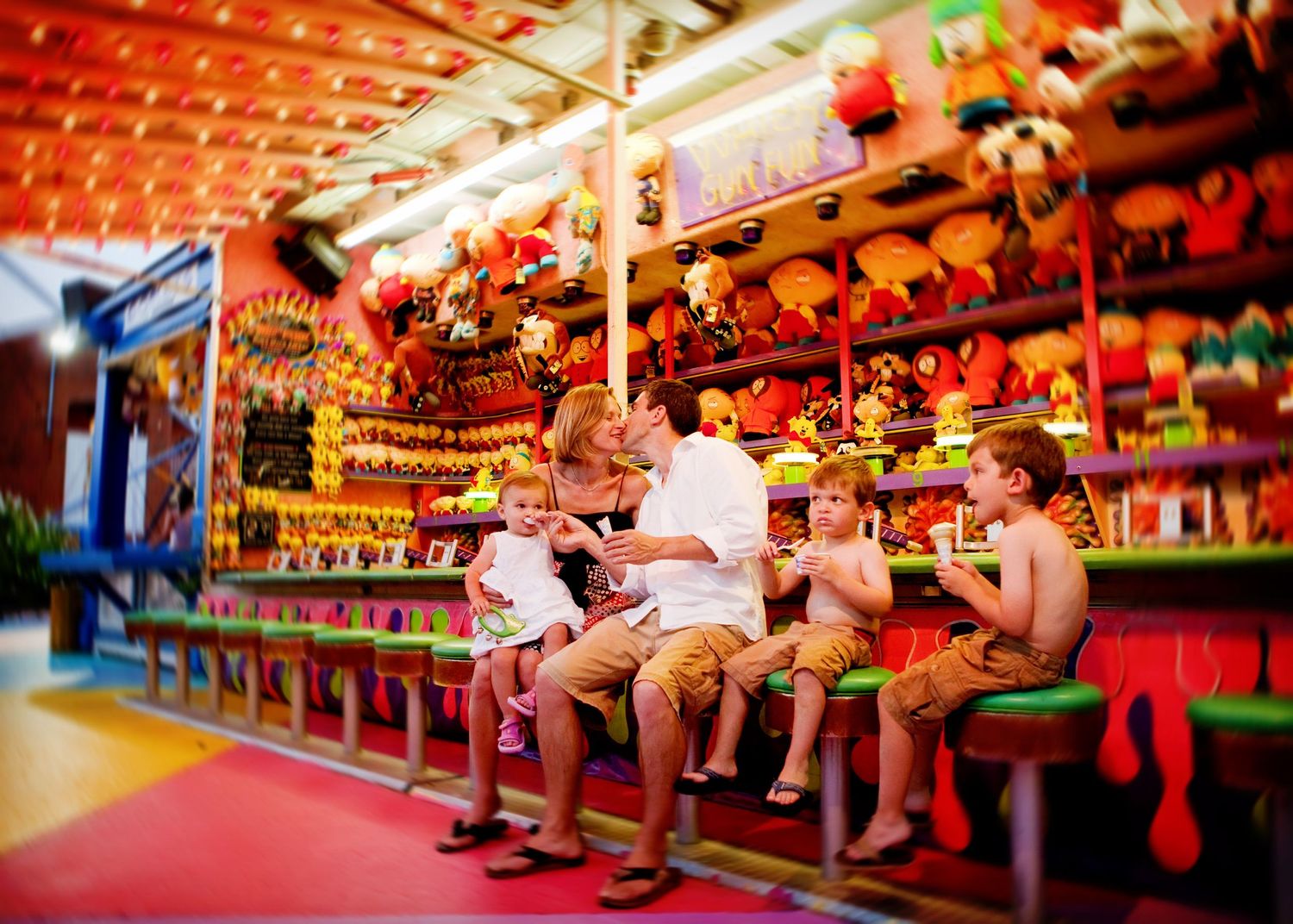 Children enjoy treats while sitting at a colorful carnival game booth filled with stuffed animal prizes.