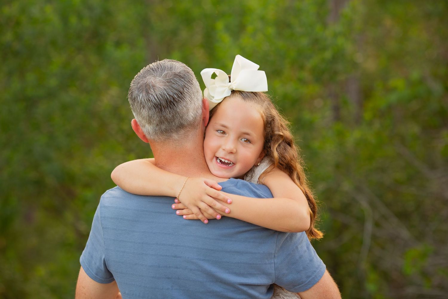 A young girl with a white bow hugs a gray-haired man outdoors, smiling joyfully against a green background.