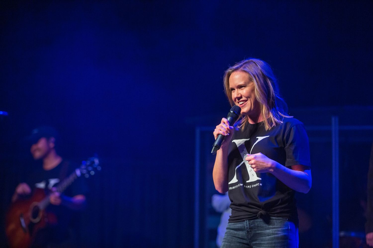 Performer singing on stage with blue lighting and guitarist in background.