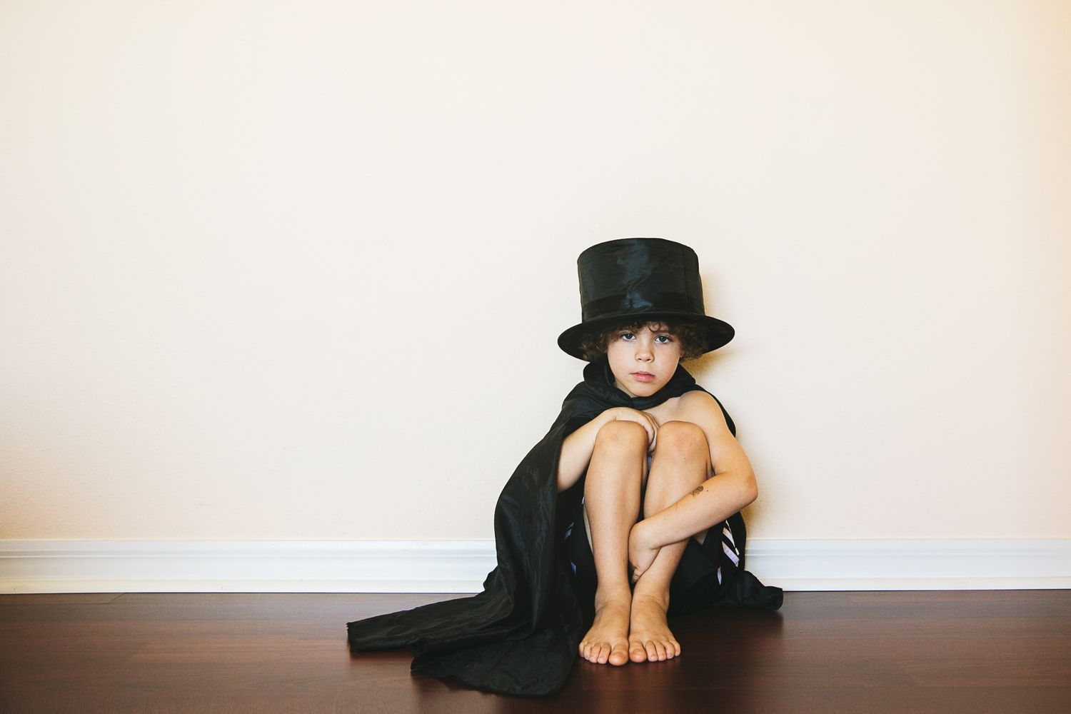 A minimalist portrait of someone wearing a black hat and coat while sitting against a white wall.