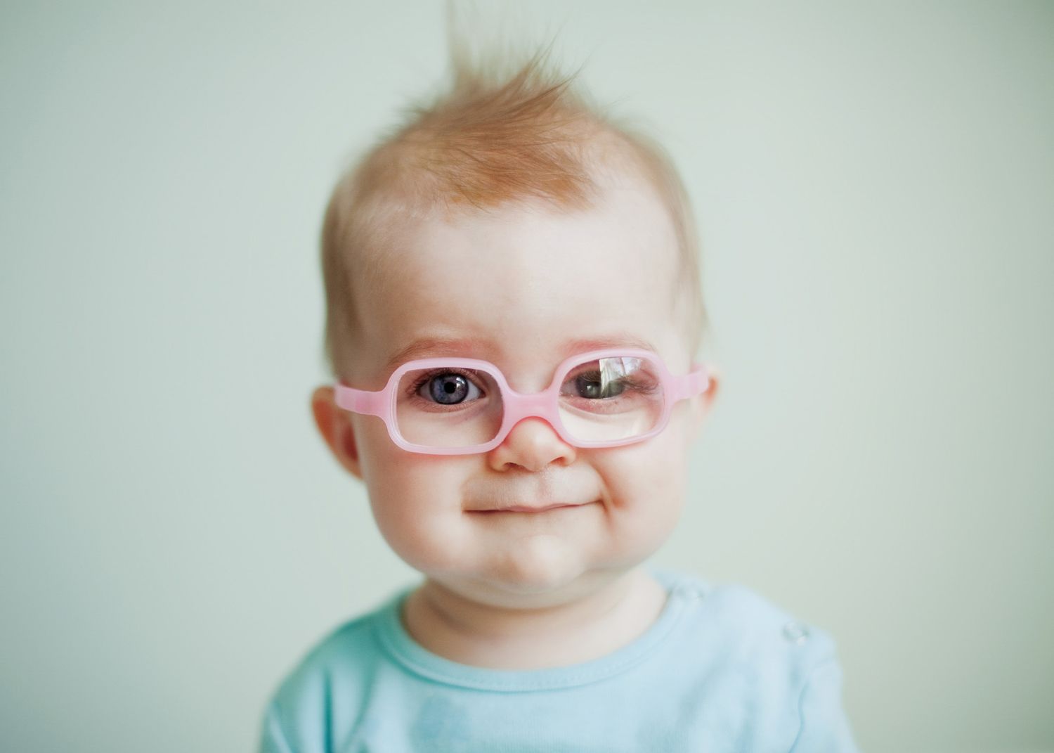 Baby wearing pink glasses smiles sweetly in close-up portrait against light background.
