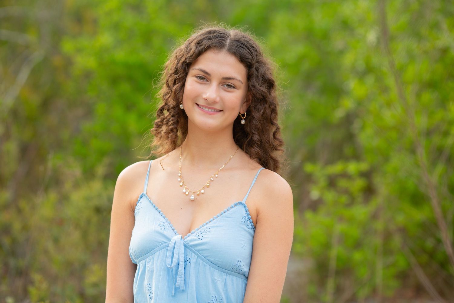 Young woman with curly hair wearing a light blue sundress and pearl necklace smiling outdoors with green foliage background.
