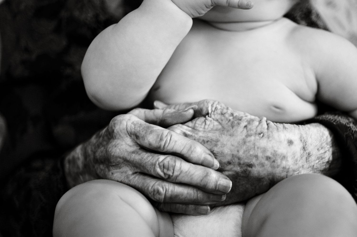 A black and white close-up of weathered hands gently holding a baby's bare skin.