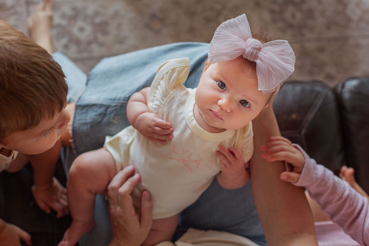 Adorable baby girl in white outfit and pink bow headband being held up by parents on a couch.