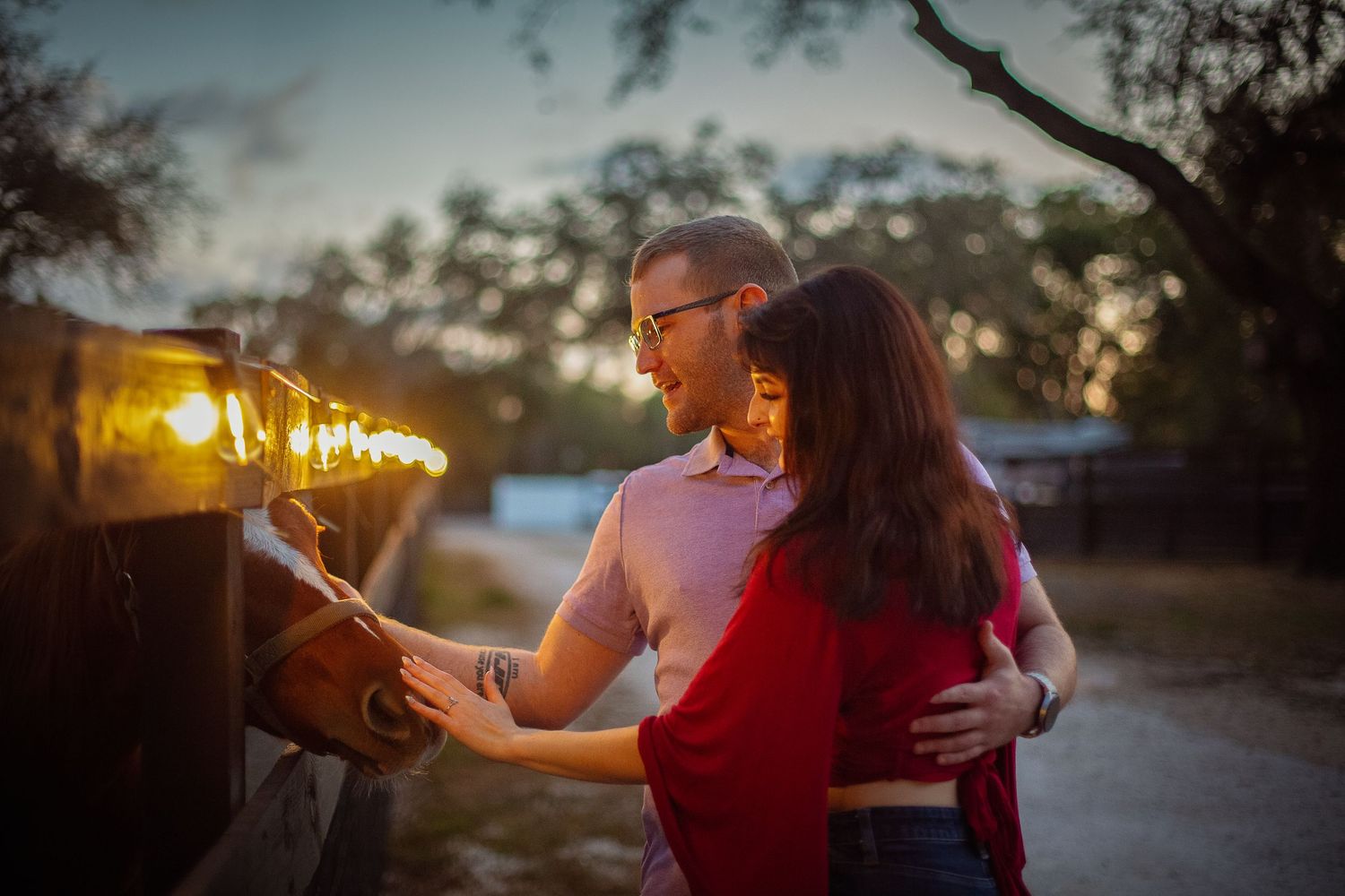 Couple embracing romantically at dusk near glowing string lights in an outdoor setting.