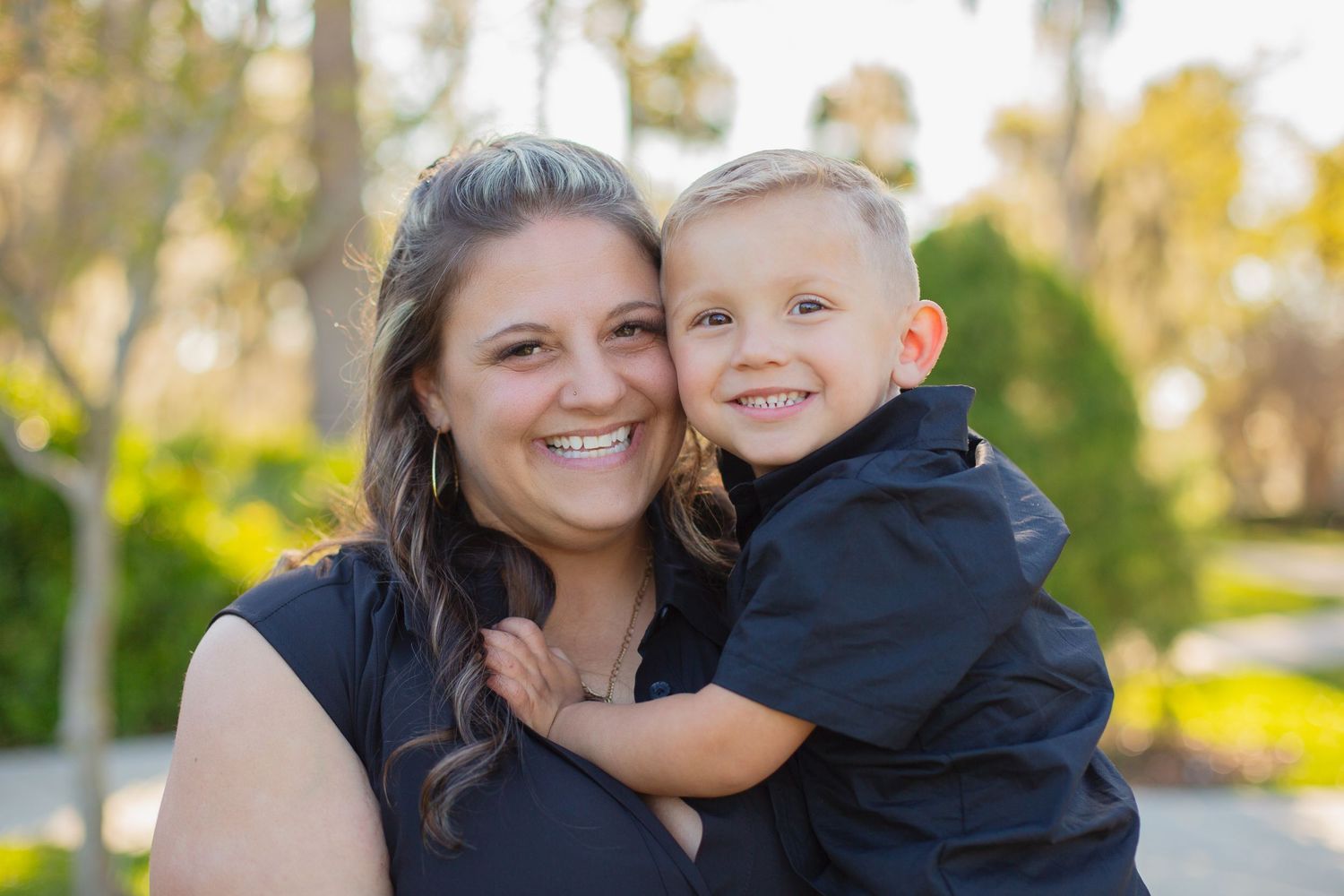 A parent wearing a black top and their young child in navy blue smile together outdoors on a sunny day.