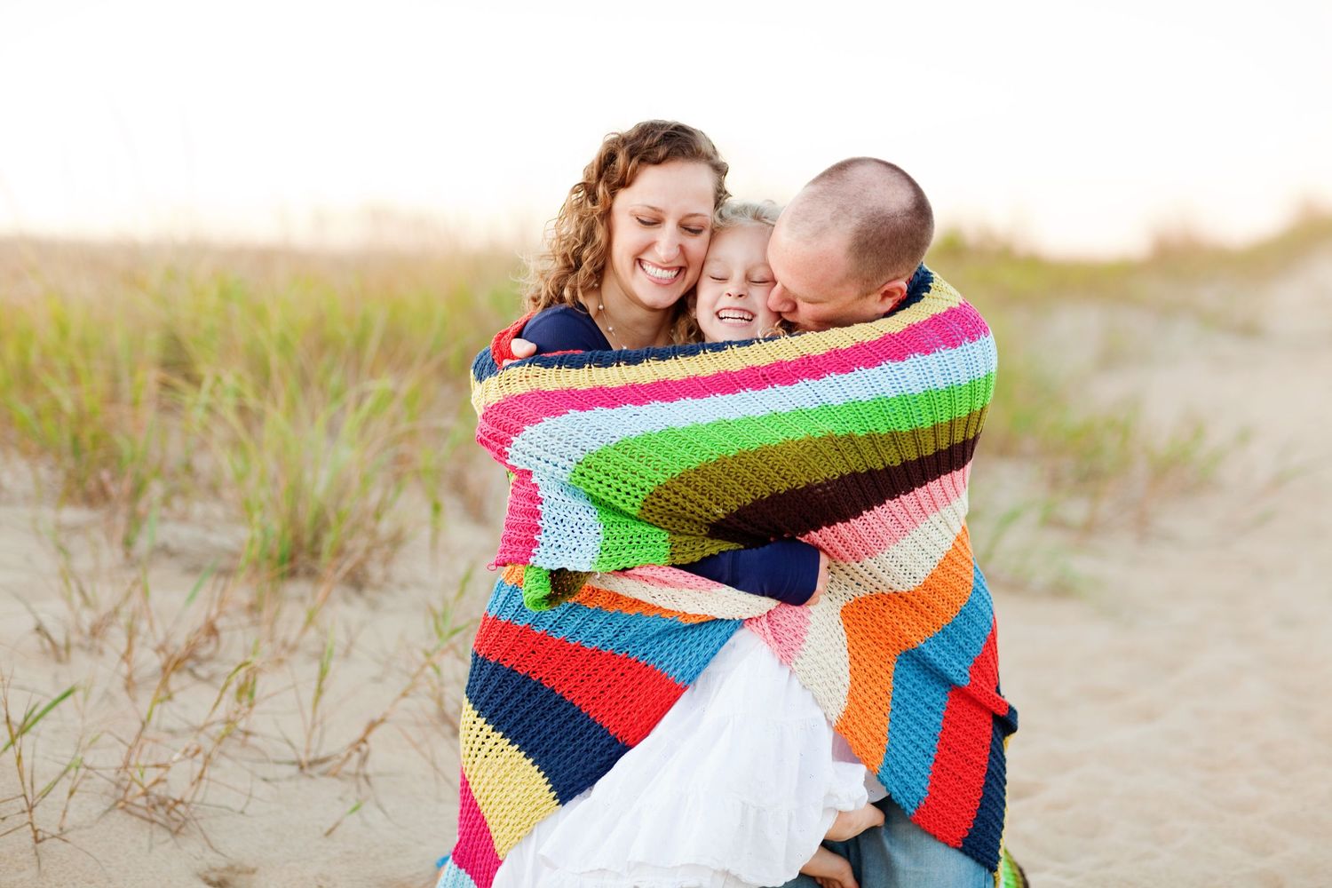 A family wrapped in a colorful striped blanket embracing on a sandy beach.