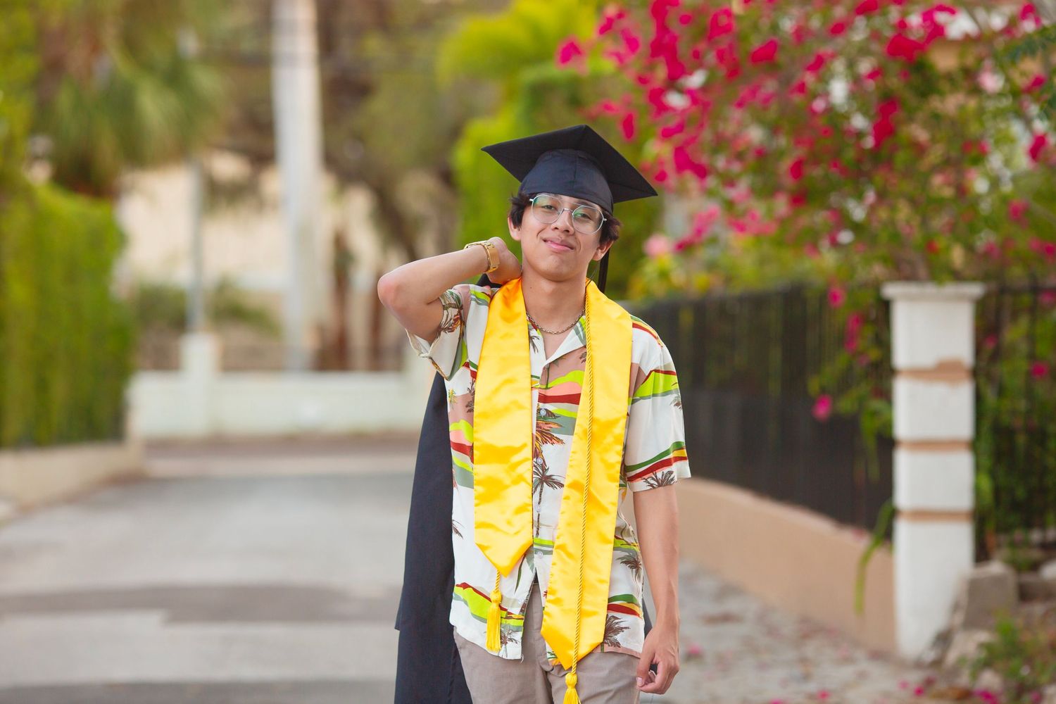 Young man in graduation cap and yellow stole smiling outdoors near pink flowers on a sunny day.