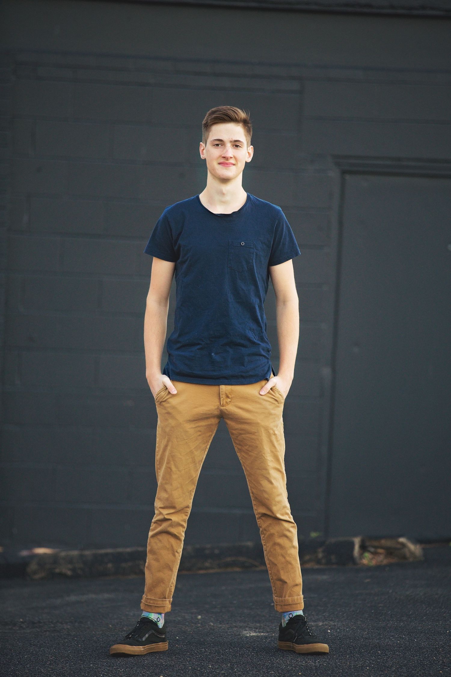 A young person in navy t-shirt and khaki pants stands casually against a dark wall.
