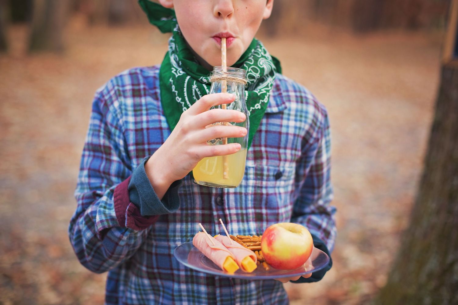 Someone in a plaid shirt and bandana sips a beverage while holding fruit outdoors.