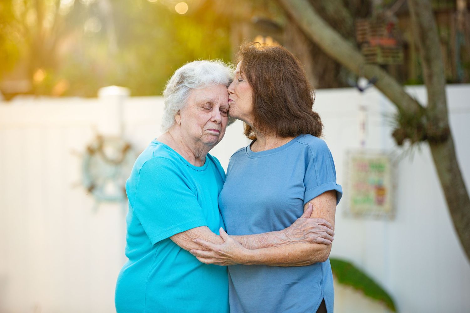 Two women in blue shirts embrace warmly in the sunlit garden.