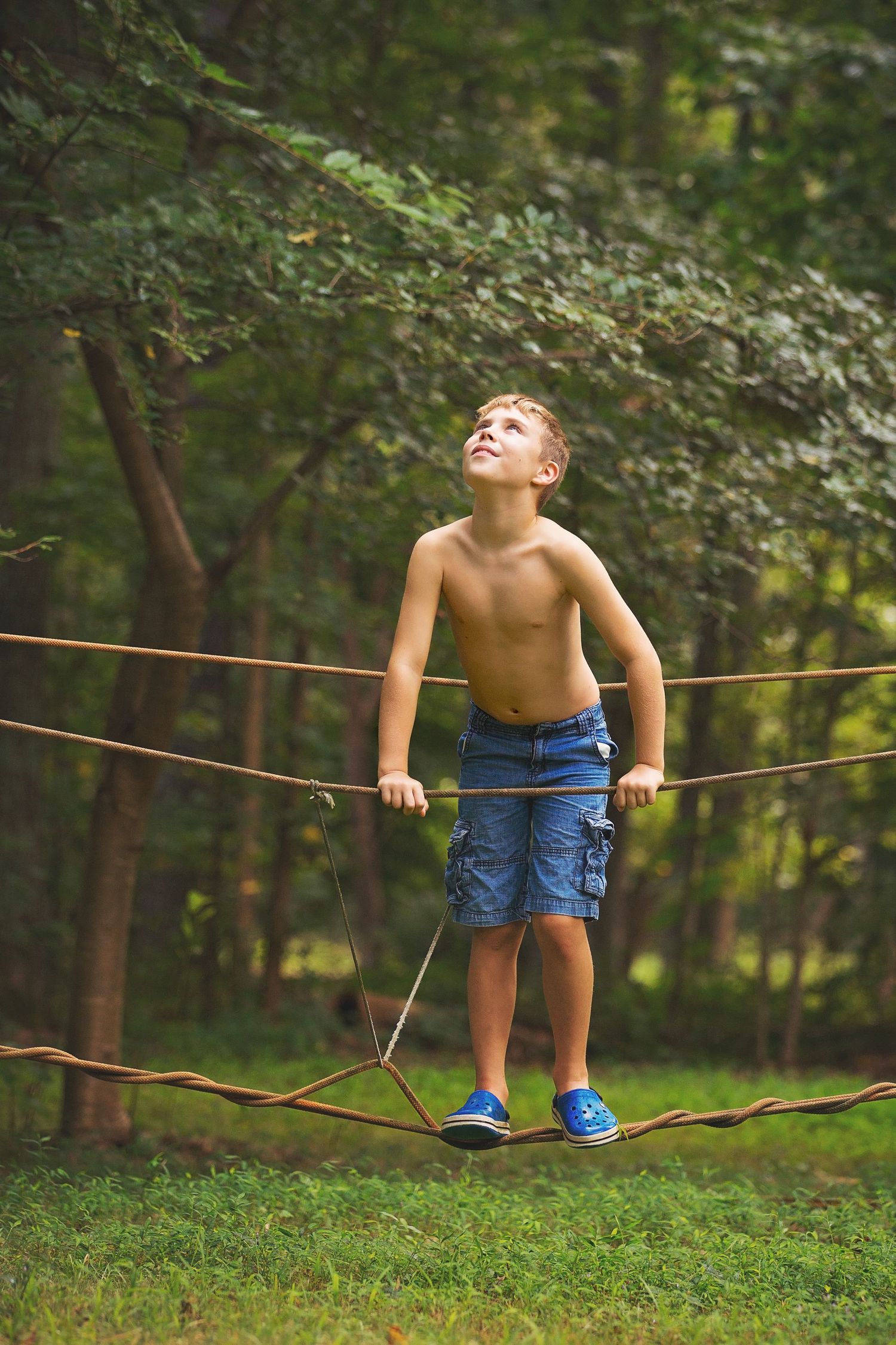 A barefoot child balances carefully on a rope bridge in a lush forest setting.