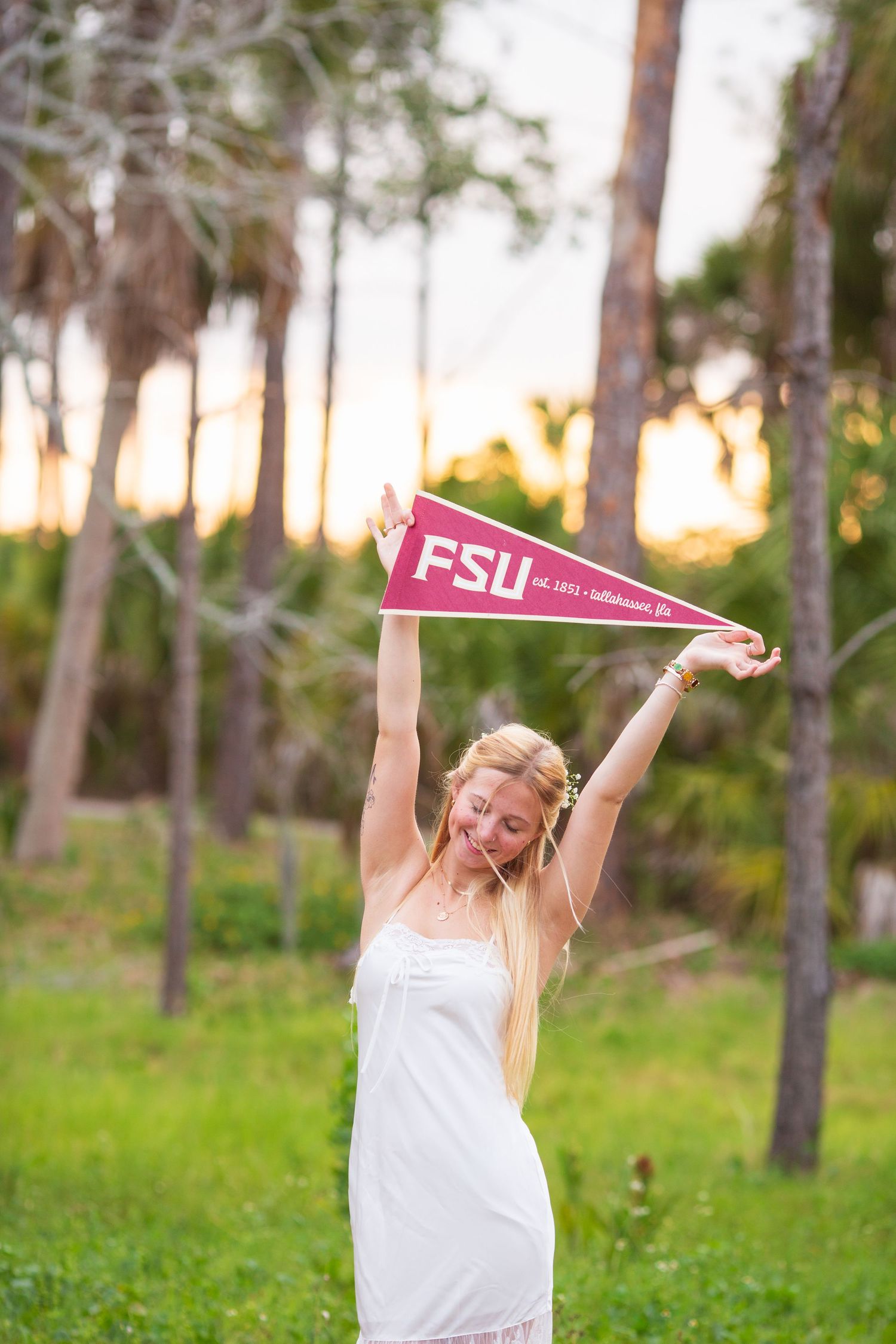 Young woman in white dress celebrating FSU acceptance, holding Florida State University pennant outdoors at sunset.