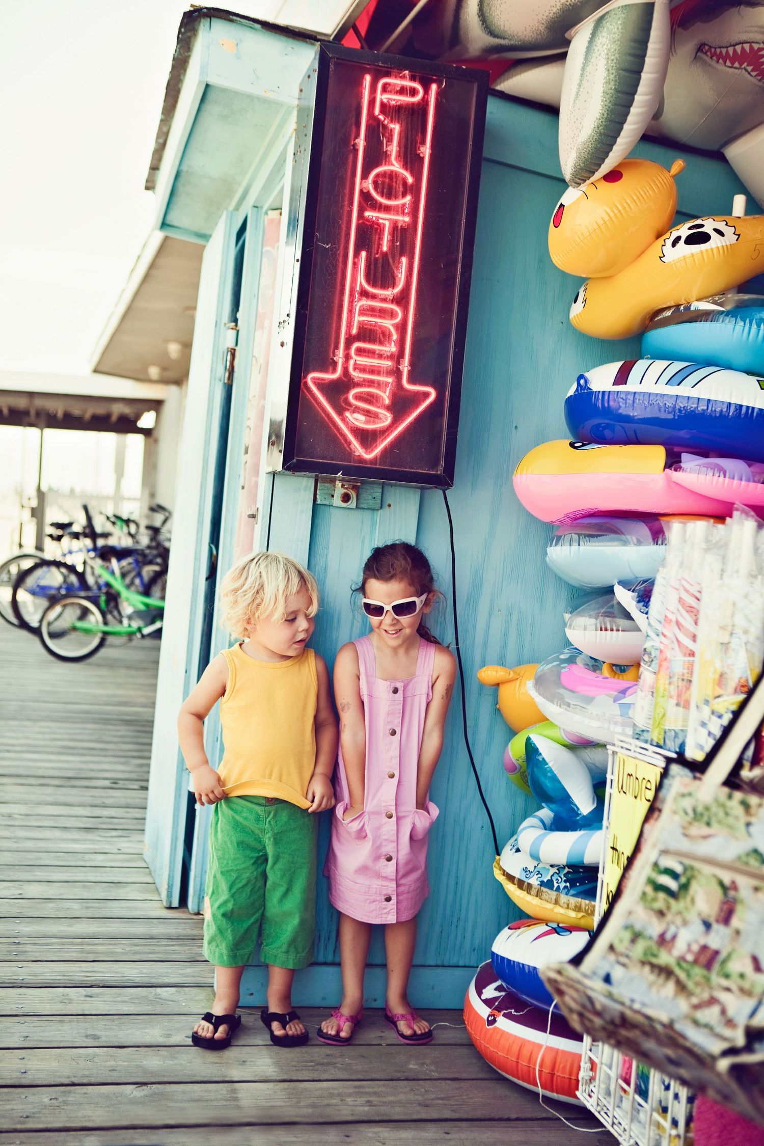 Two young people in colorful outfits standing by beach shop with pool floats and neon sign.