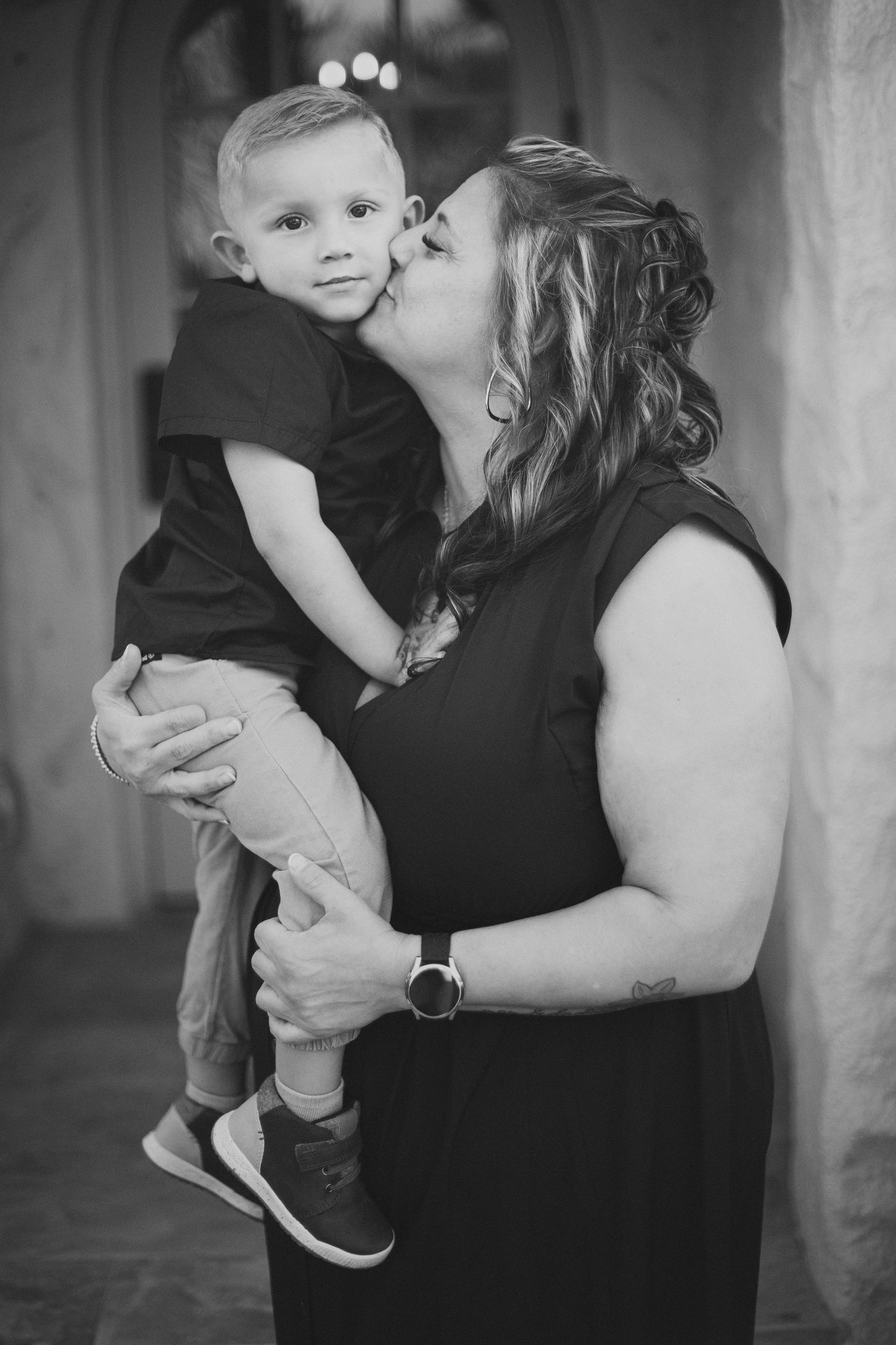 A tender black and white portrait shows a mother in a sleeveless dress lovingly kissing her small child.