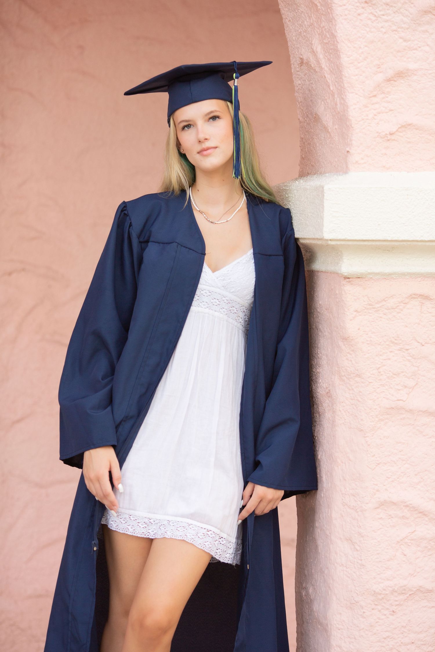 Young woman in navy blue graduation cap and gown poses against a pink stucco wall wearing a white dress.
