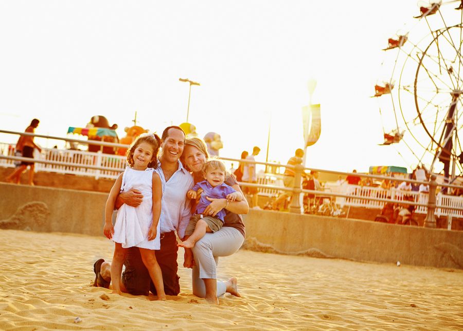 A happy family poses together at a carnival at sunset with rides visible in the background.