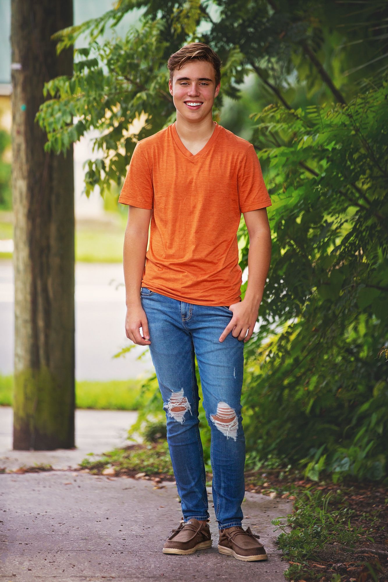 Casual portrait of person in orange v-neck and distressed jeans standing on garden path.