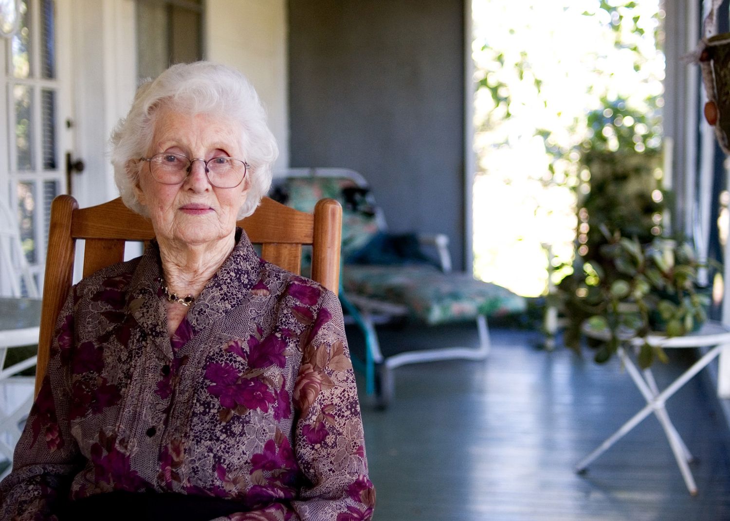 Senior woman in purple floral dress sitting on wooden porch chair.