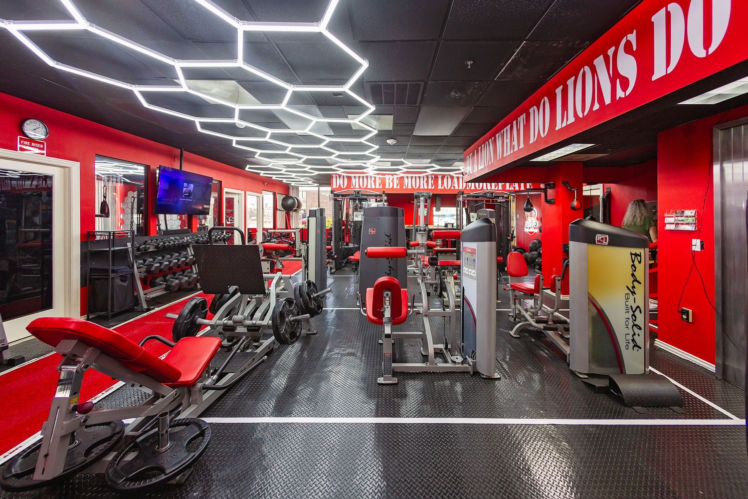 Modern red and black gym interior with weight machines and hexagonal ceiling lights.