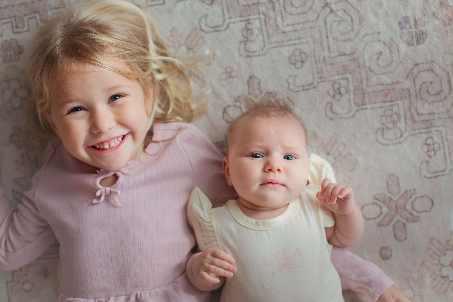 Two young sisters lying on a patterned rug, older blonde toddler smiling beside her baby sibling in white onesie.