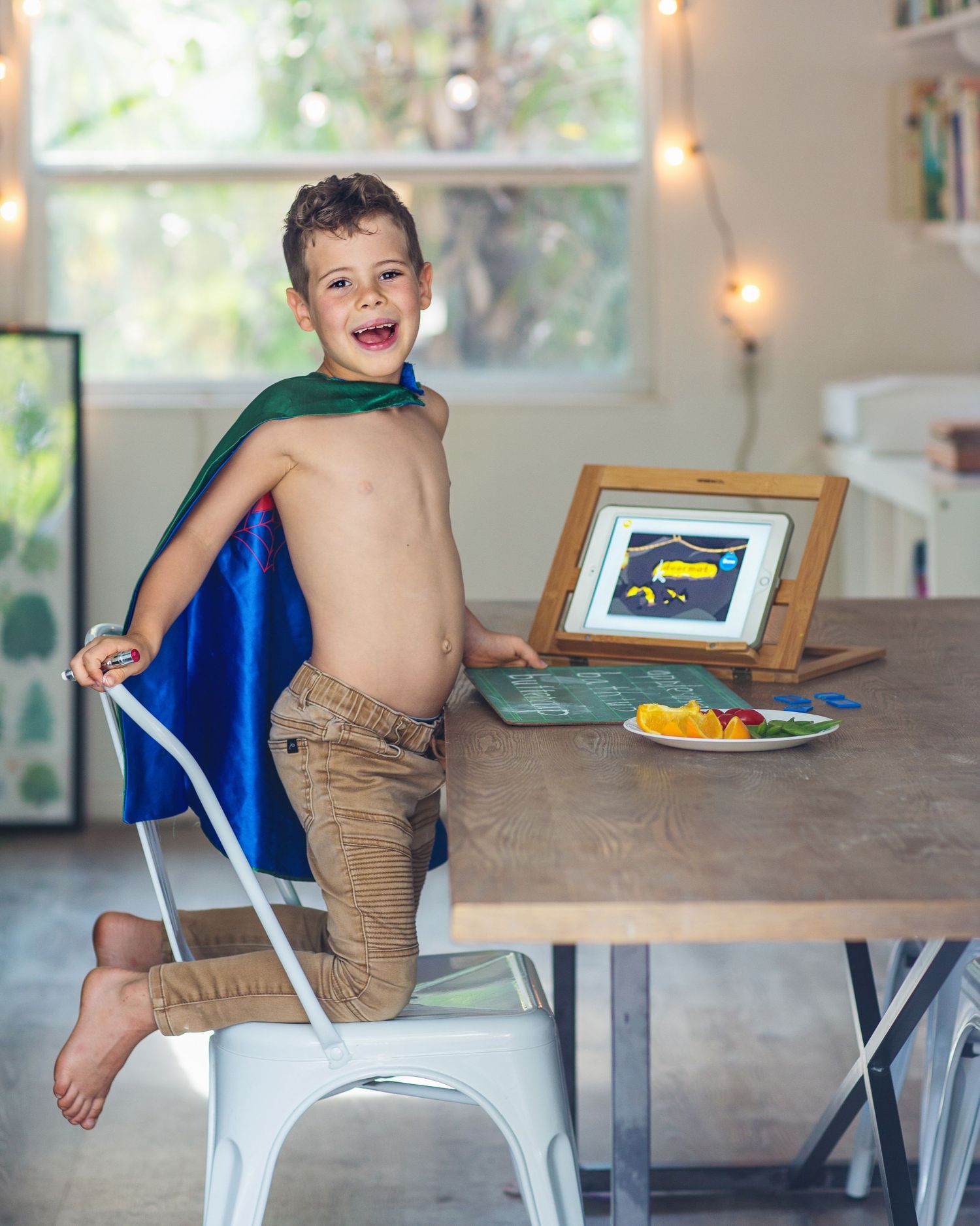 Young child in blue cape standing playfully on white chair at wooden table with plate.