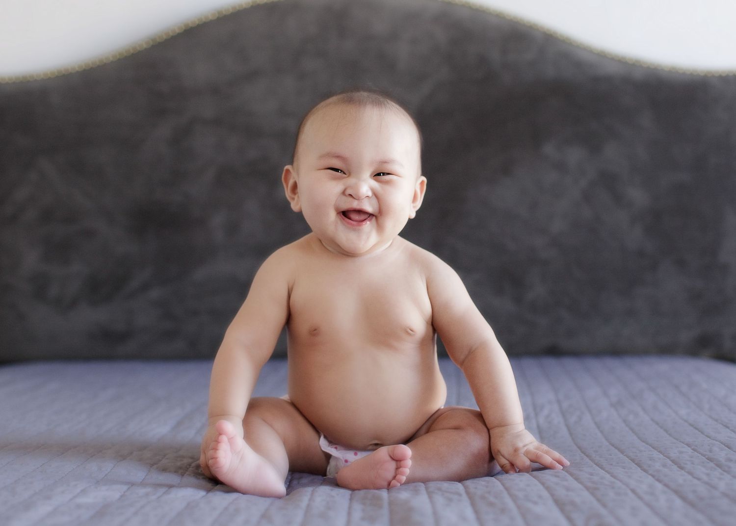 Happy baby sitting on gray bed with bright smile.