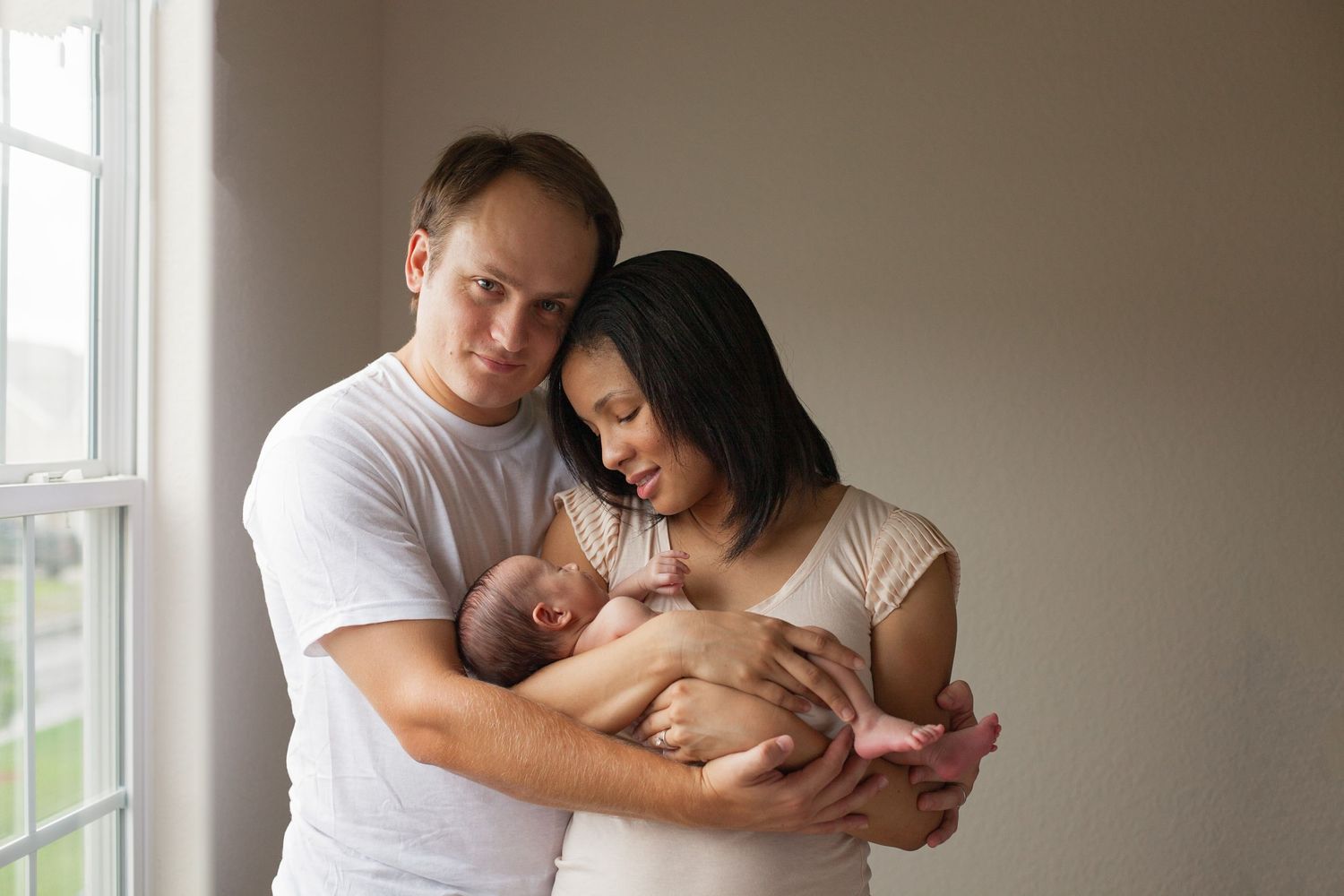 Intimate family portrait showing parents cradling newborn baby near window.