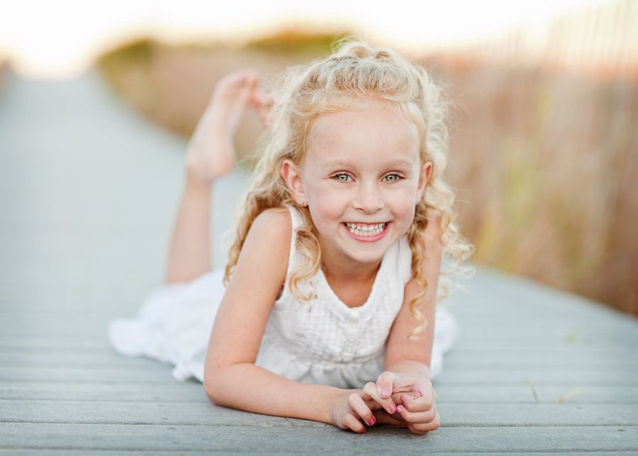 A young child in a white dress lies on a wooden deck outdoors.