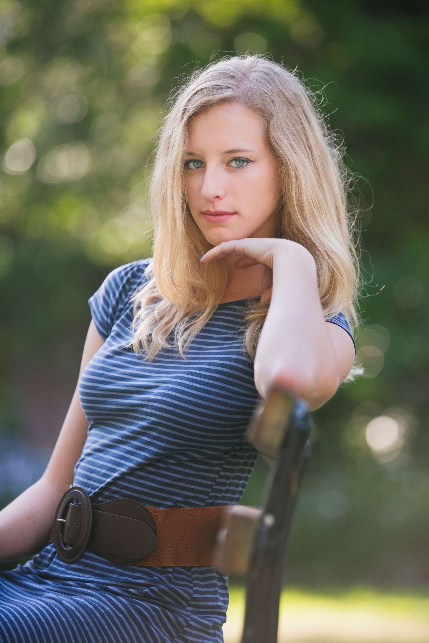 Portrait in a striped dress with brown belt sitting on a bench in a sunny garden setting.