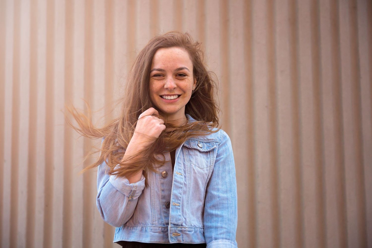 A person in denim jacket with windswept hair poses against a corrugated metal background.