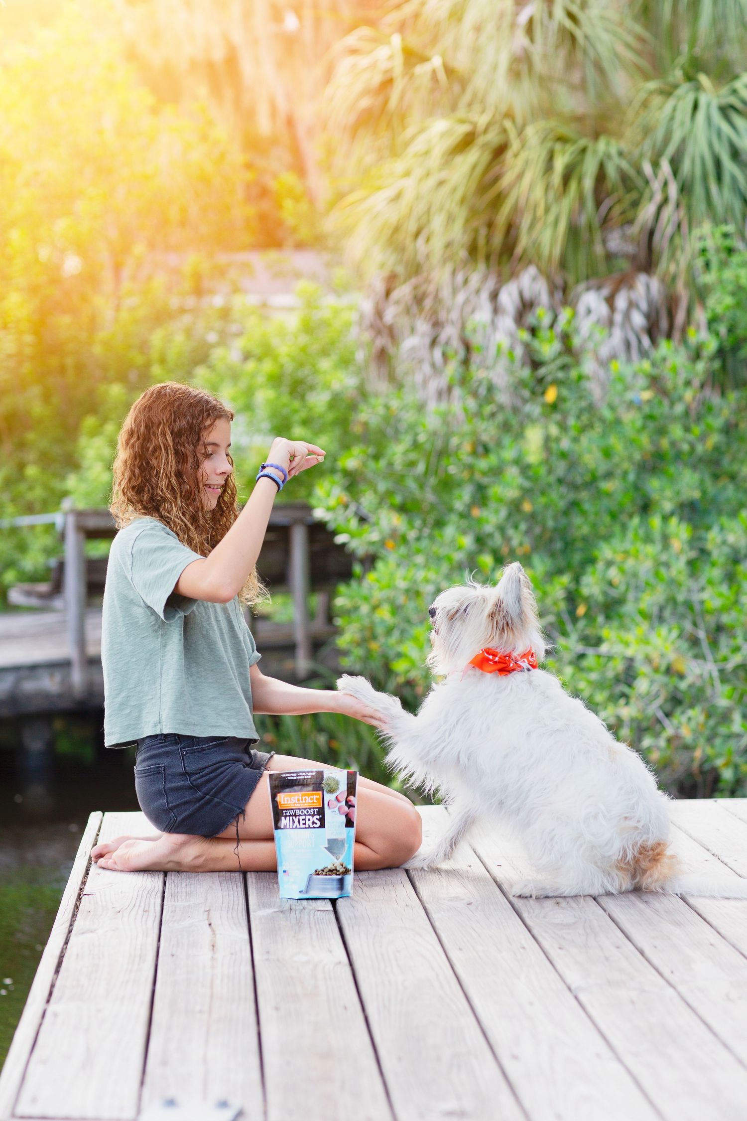 A young person sits on a dock with a white dog enjoying snacks on a sunny afternoon.