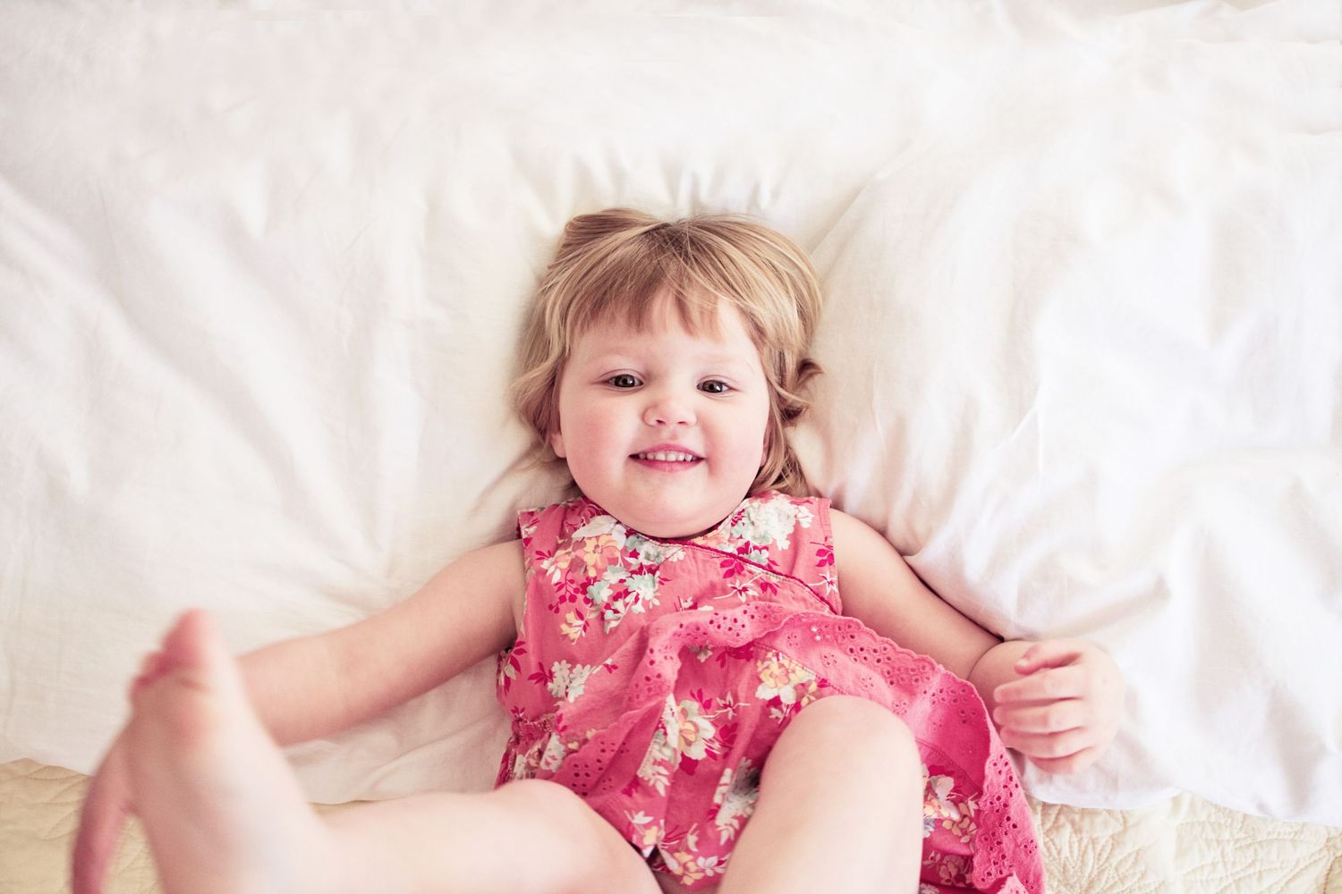 A joyful toddler wearing a pink floral dress laying on white bedding.