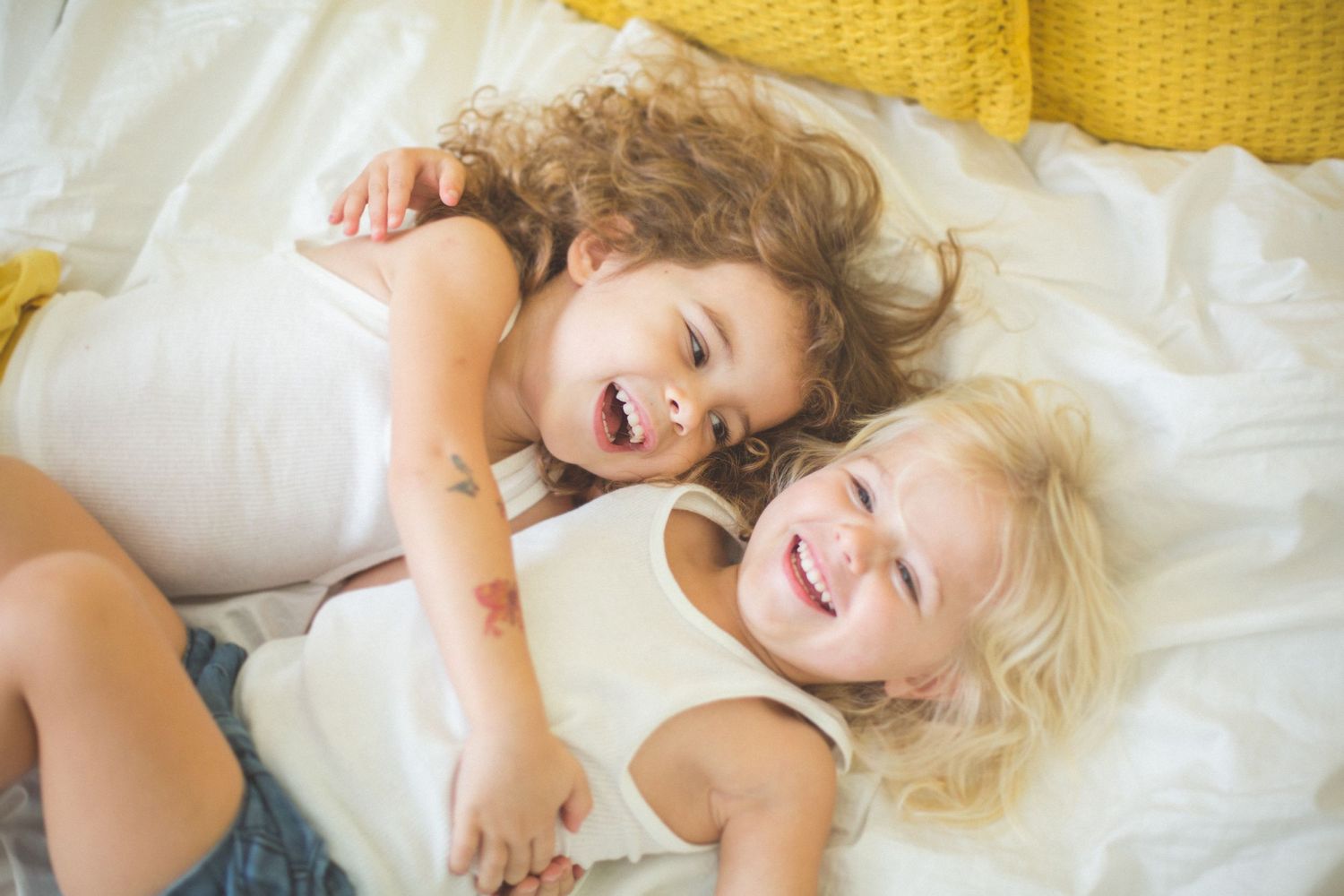 Two children lying on a white bed laughing and playing together.