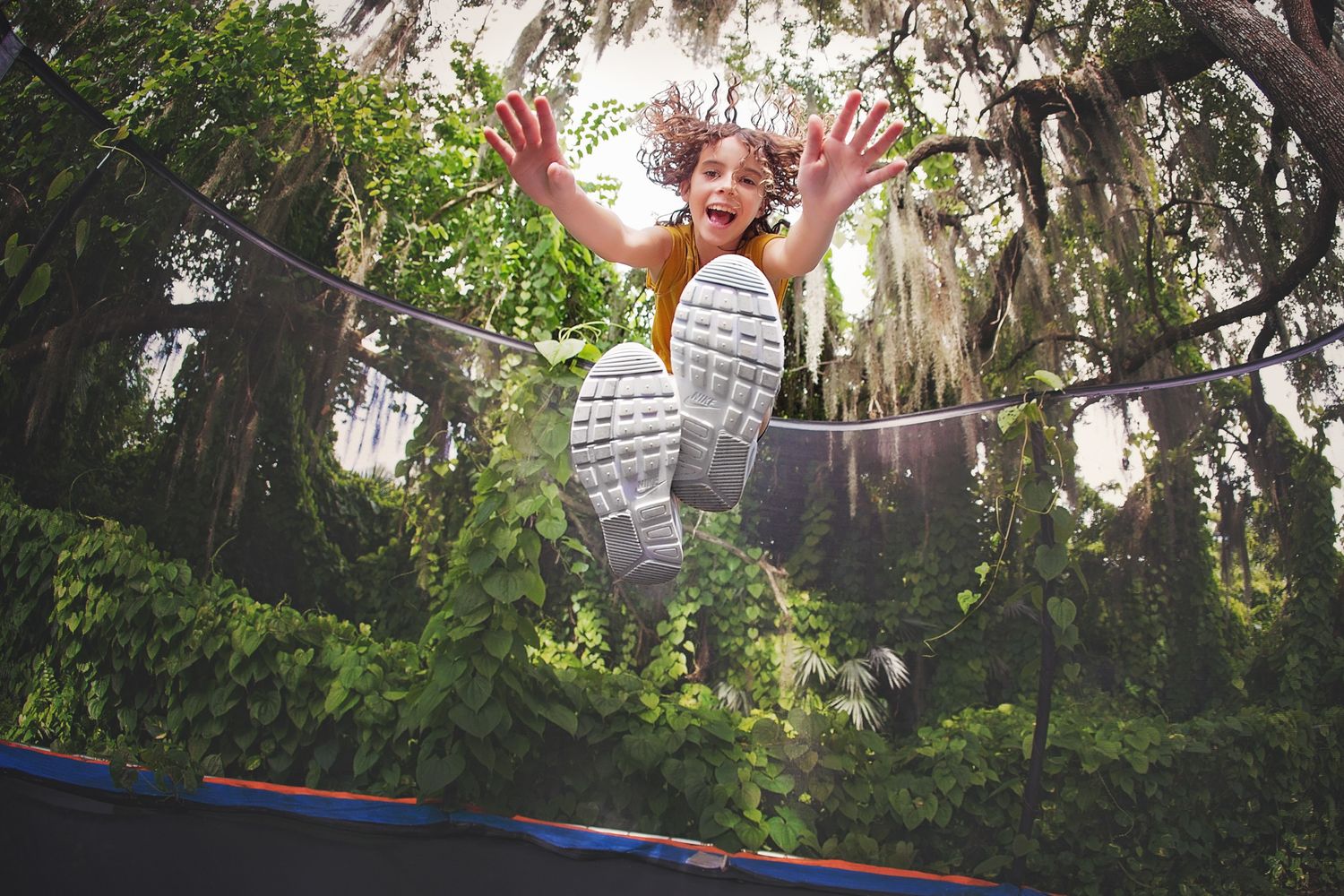 Child jumping high on backyard trampoline with mossy trees in background.
