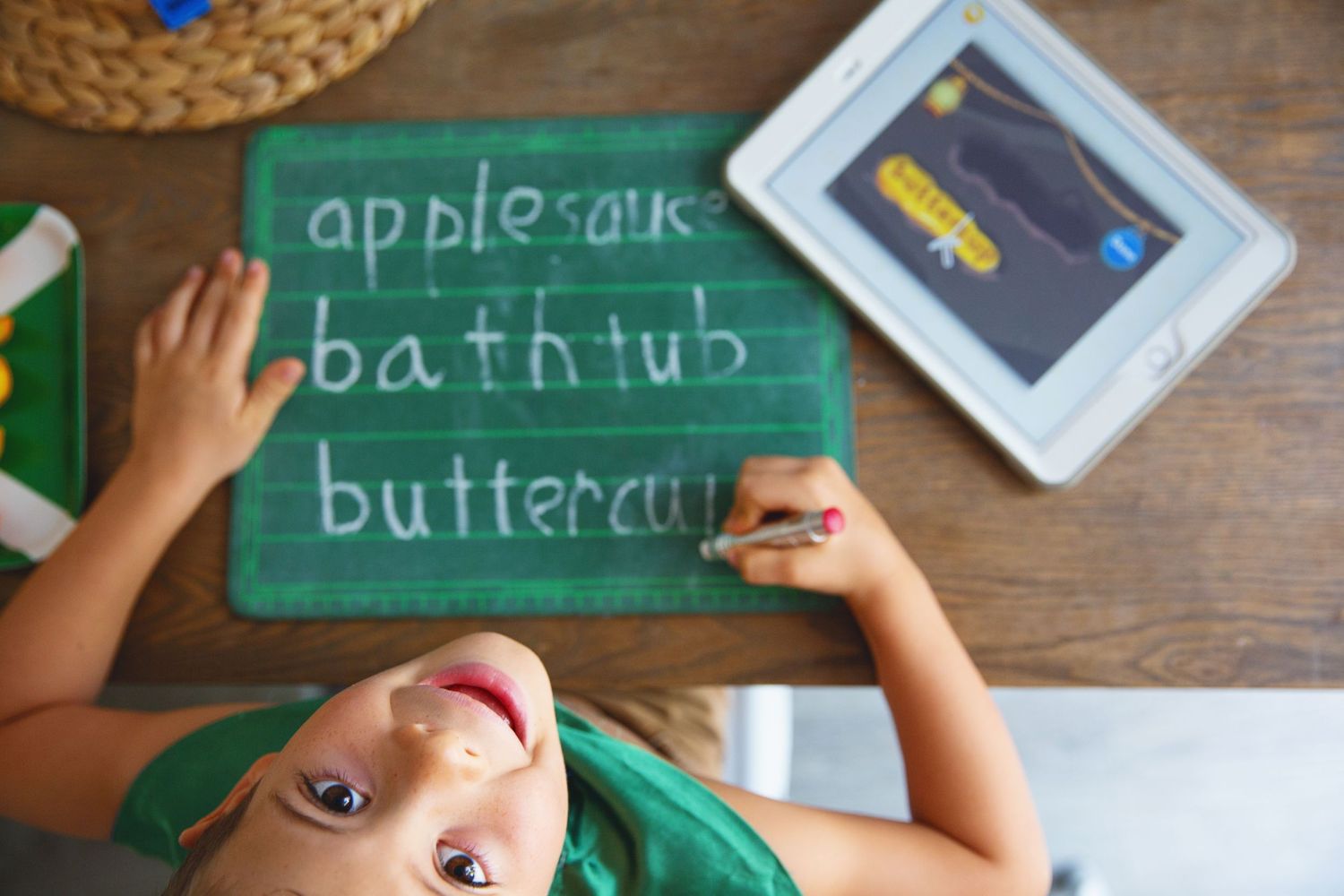 Student practicing handwriting on a green chalkboard with tablet nearby.
