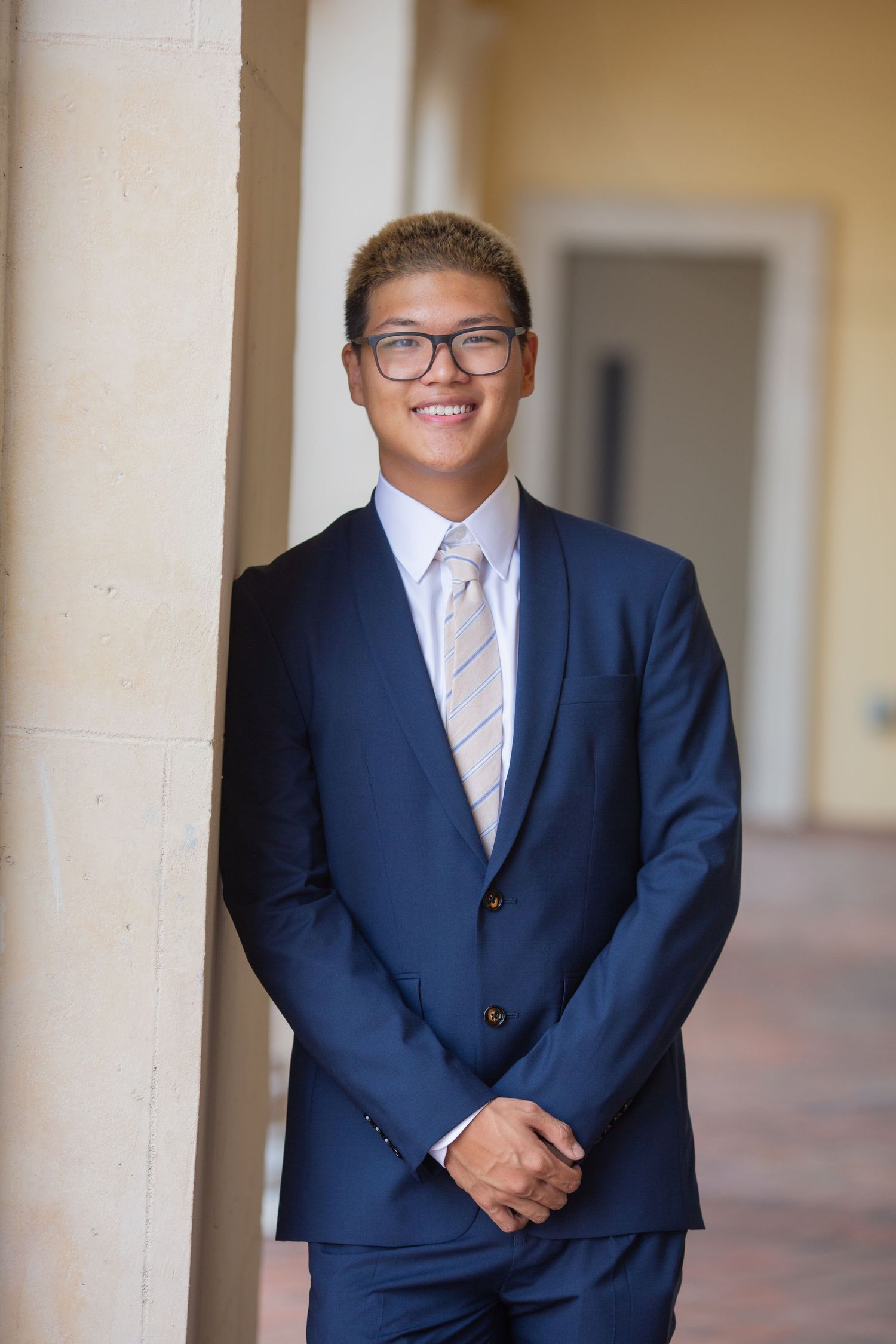 Young man in navy blue suit and tie with glasses smiling confidently while leaning against a column outdoors.