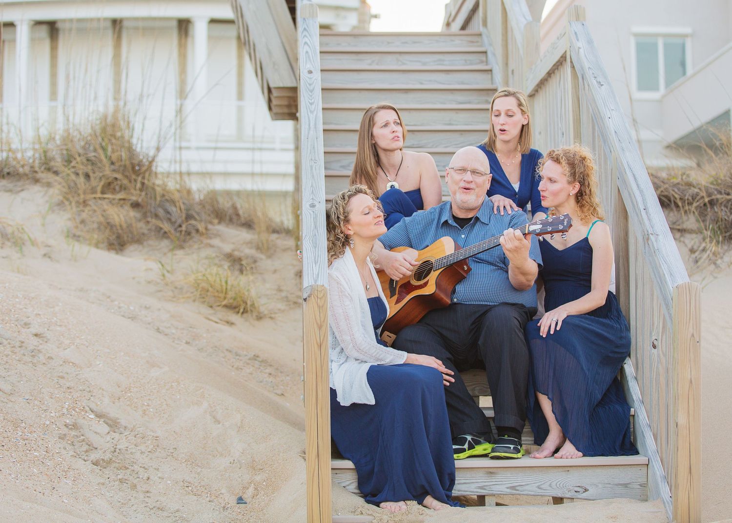 A family gathers on beach house stairs while someone plays guitar.