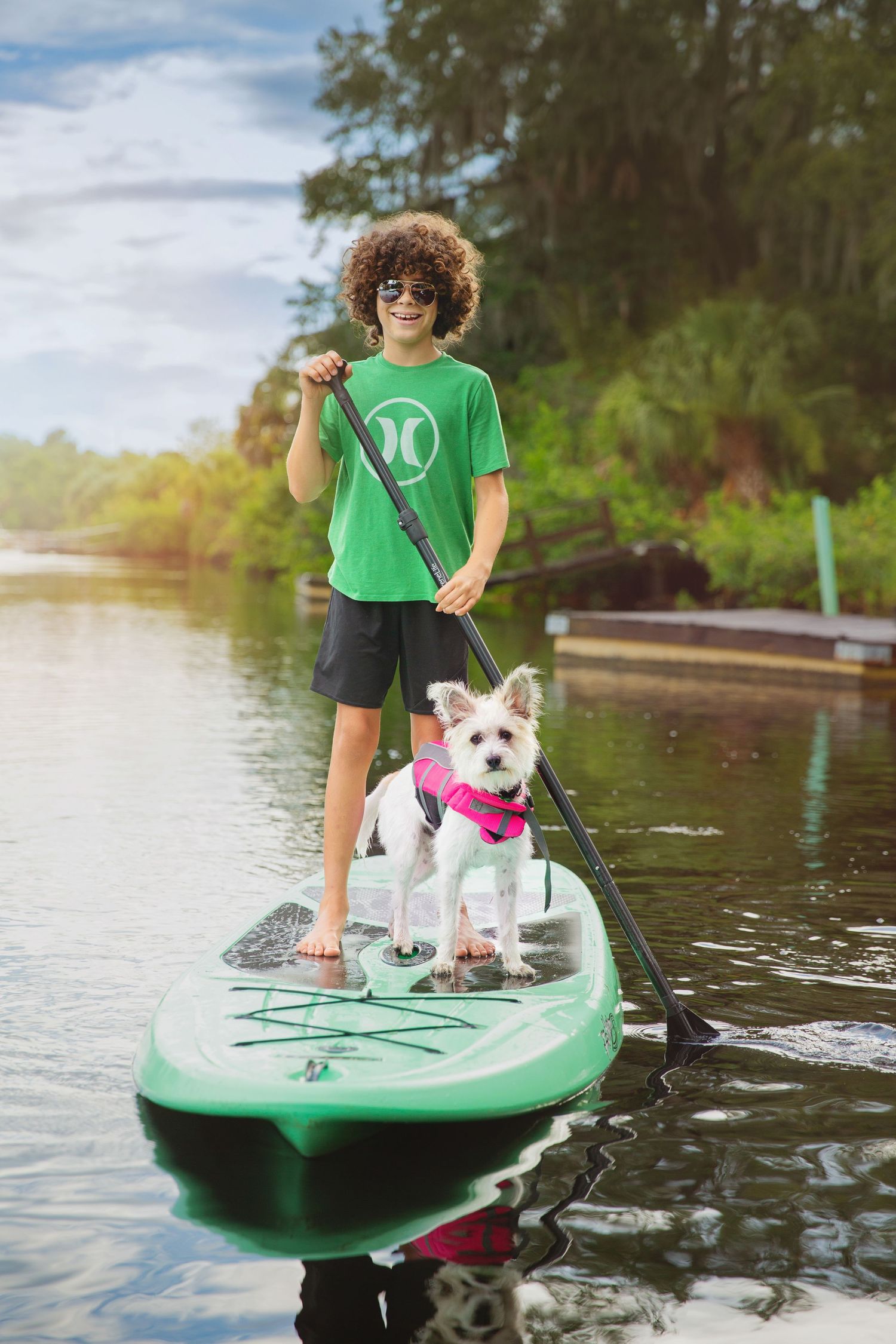 Person paddle boarding with a small white dog wearing a pink vest on calm waters.
