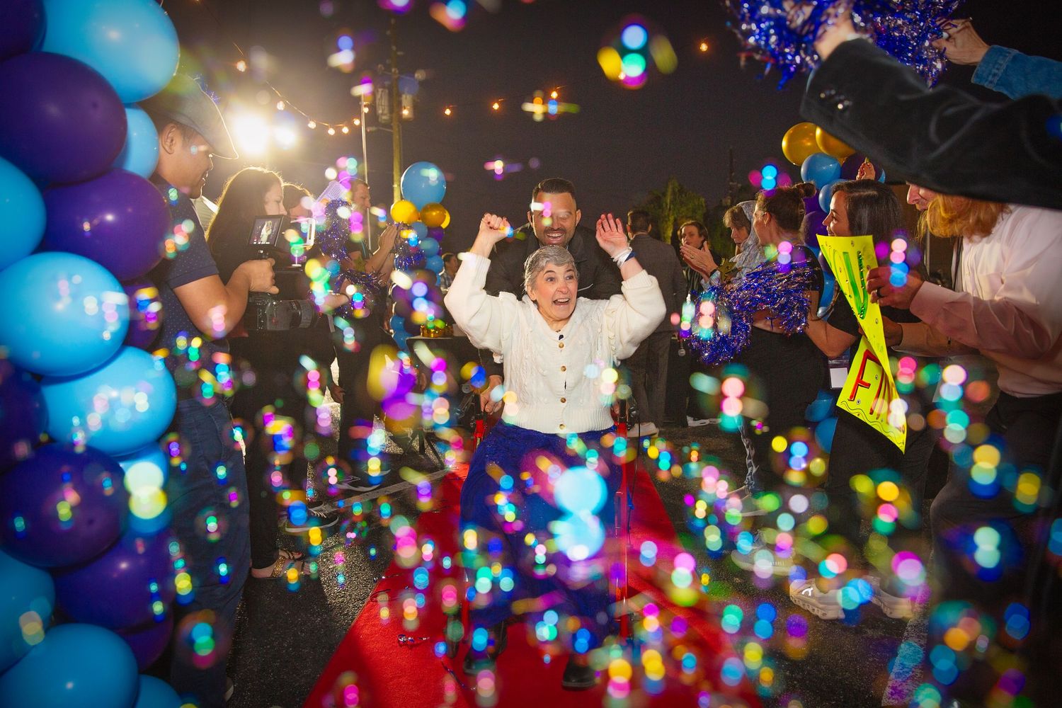 Woman celebrates with raised arms on red carpet surrounded by colorful balloons and bokeh lights at night event.