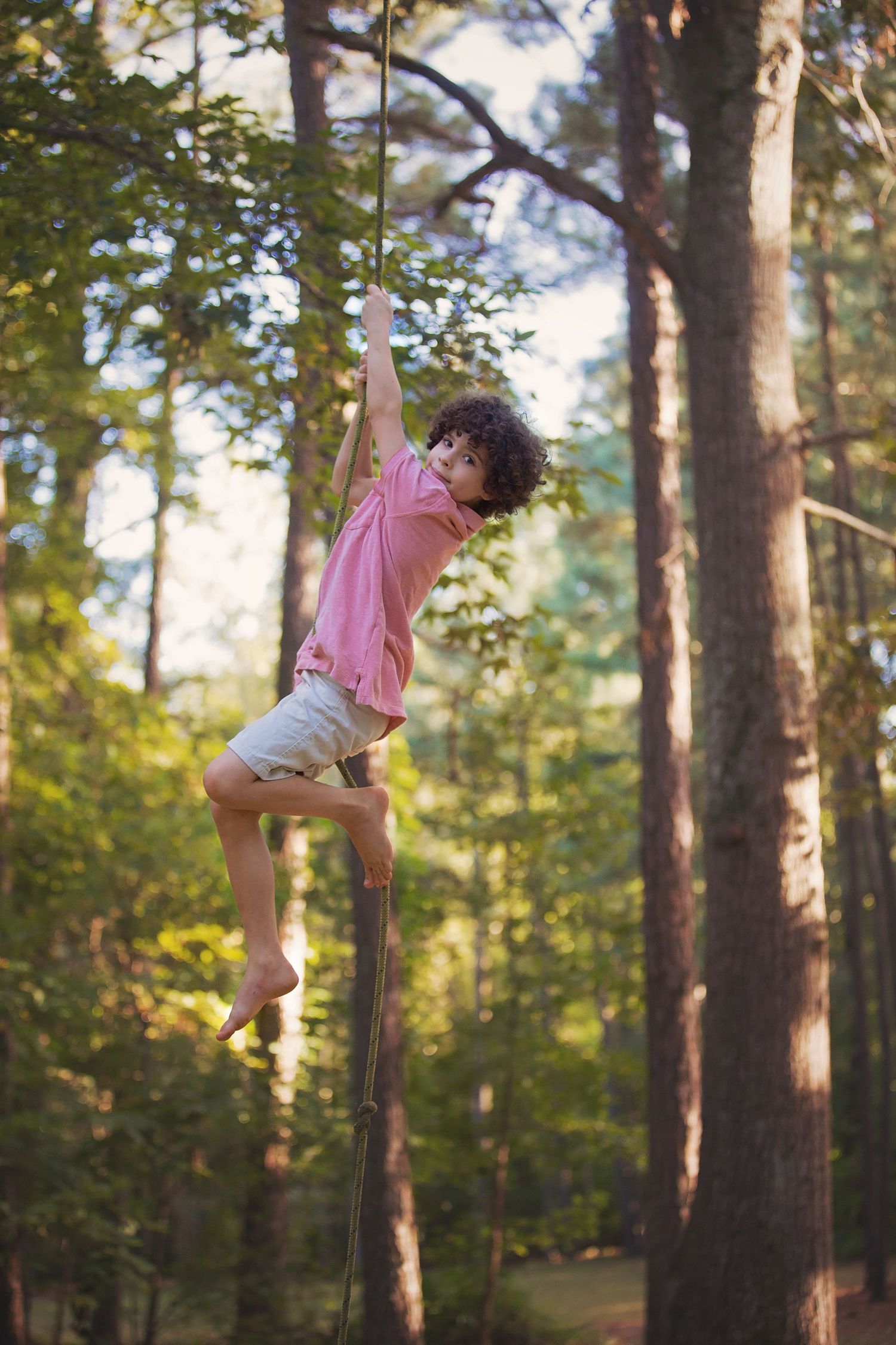 Child climbing high on pole in pink shirt against forest backdrop during golden hour.
