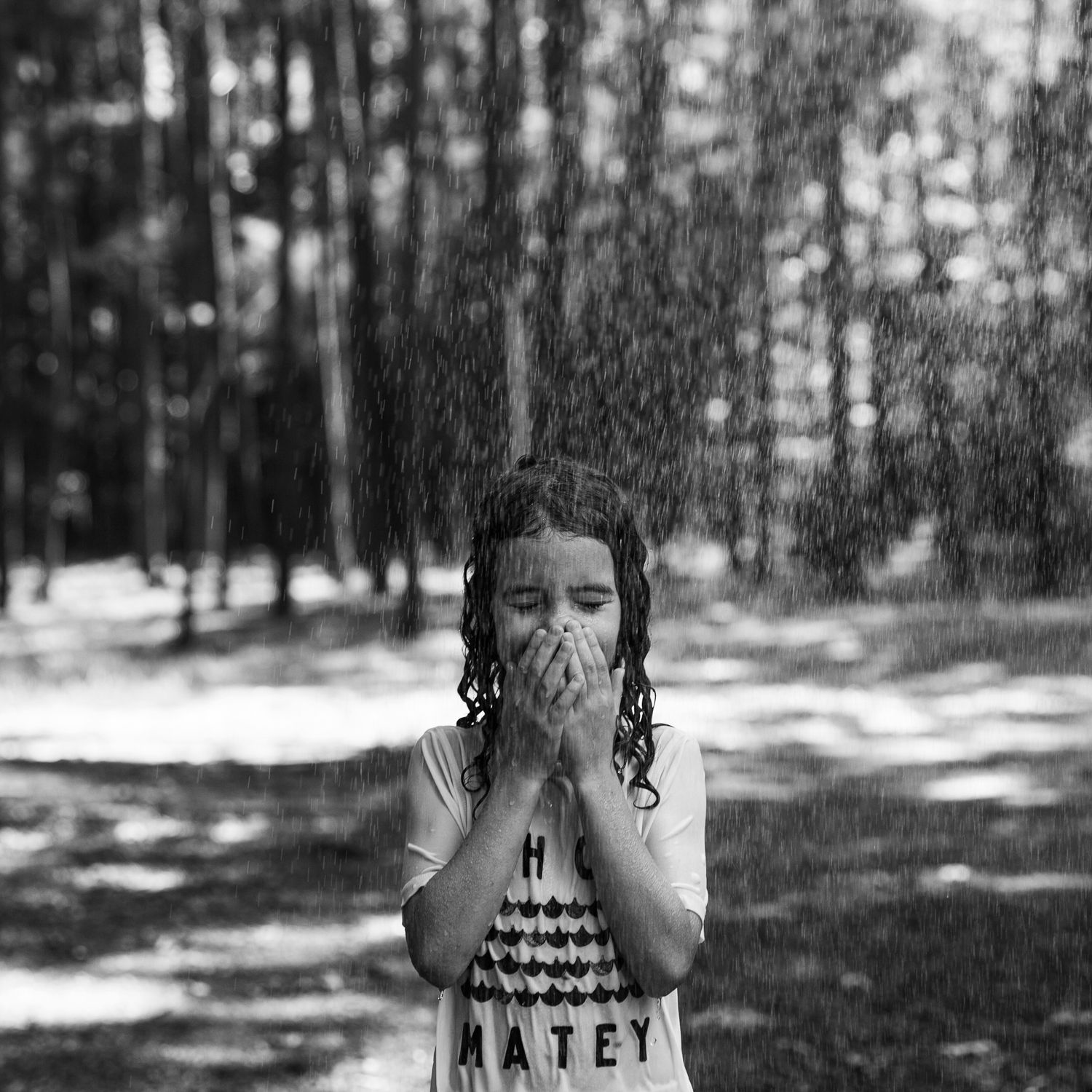 A child standing in dappled sunlight with hands covering face in a wooded setting.
