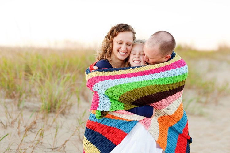 Family wrapped together in a colorful striped blanket at the beach during sunset.