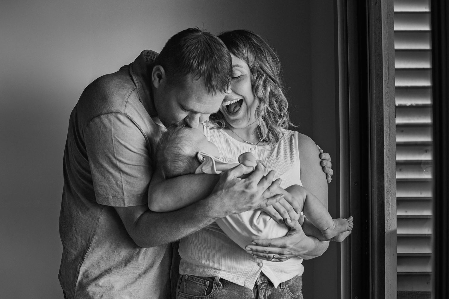 Black and white photo of parents tenderly kissing their newborn baby by a window with shutters.