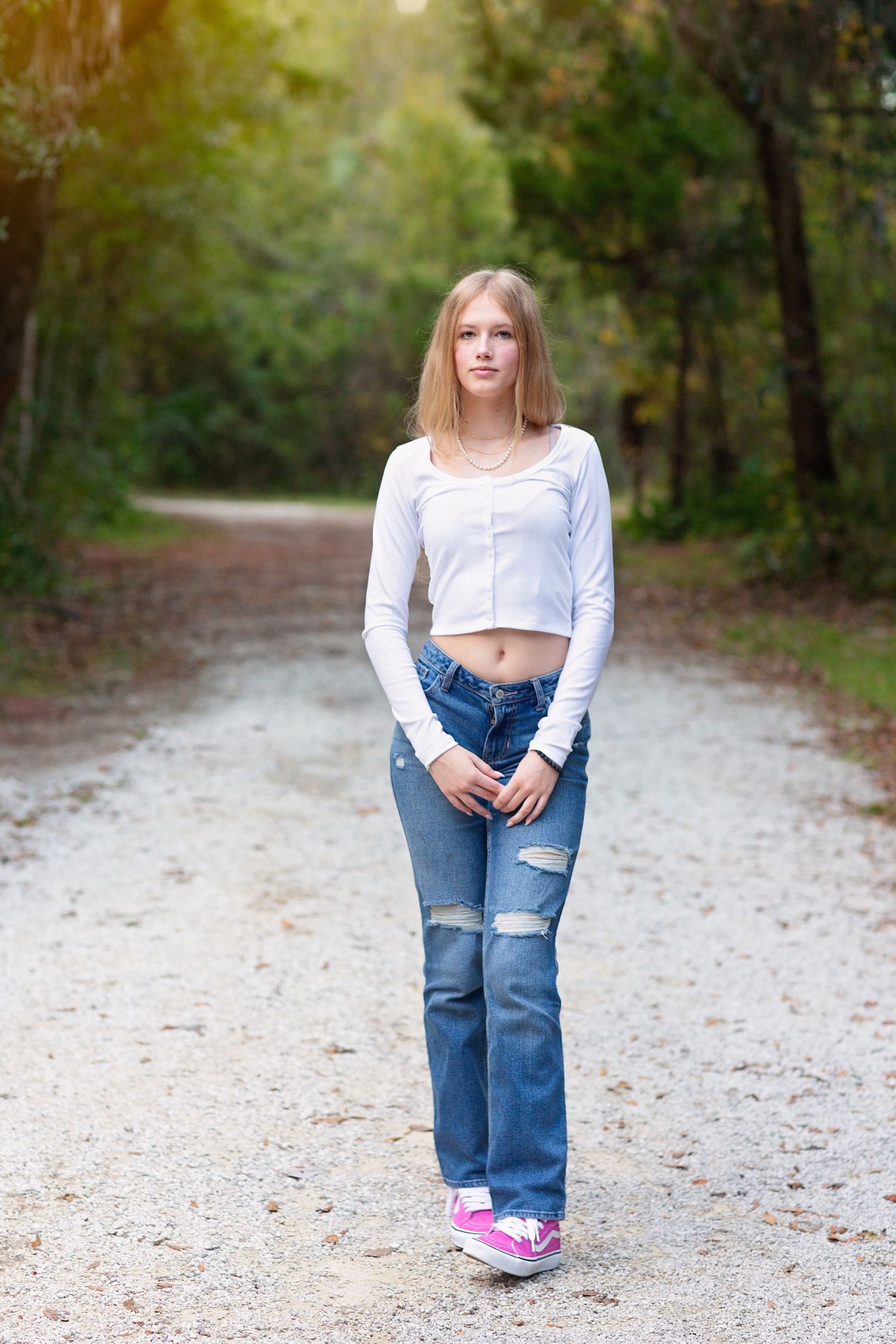 Person in casual attire standing on a wooded path wearing ripped jeans and white crop top.