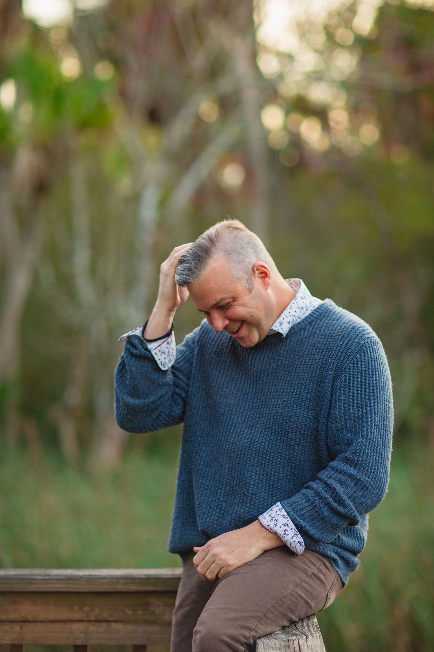A person in a blue sweater laughs while sitting on a wooden surface outdoors.