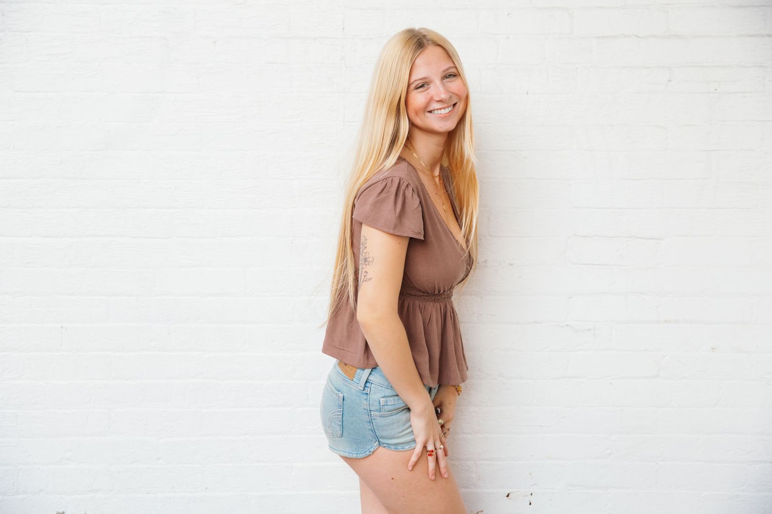 Smiling blonde woman in brown top and denim shorts posing against white brick wall.